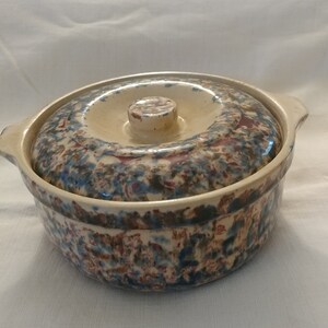 Può includere: Una casseruola in ceramica vintage con coperchio. La casseruola è decorata con un motivo maculato blu, marrone e rosso. Il coperchio ha un pomo in cima.