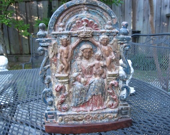 May include: A rectangular, arched relief plaque featuring a central figure holding a child, flanked by two winged cherubs placing a crown above their heads. The piece is finished in distressed blue, red, and gold paint, mounted on a dark wood base.
