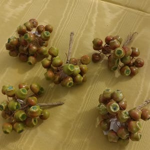 May include: Five clusters of artificial berries with green and brown hues. Each cluster has multiple berries attached to a small twig. The berries have a glossy finish and are arranged on a yellow surface.
