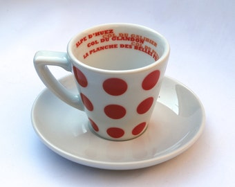 May include: White ceramic coffee cup with red polka dots and the text "Alpe d'Huez, Col du Galibier, Col du Glandon, La Planche des Belles Filles" printed around the rim.