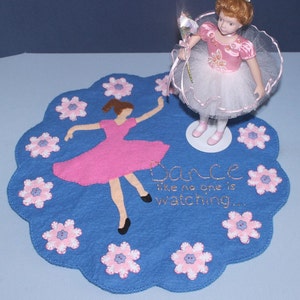 Op de afbeelding: Een blauwe vilten placemat met een roze ballerina-ontwerp en de geborduurde tekst "Dance like no one is watching...". Een ballerina pop in een roze en witte tutu staat op de mat, omringd door roze bloemaccenten.