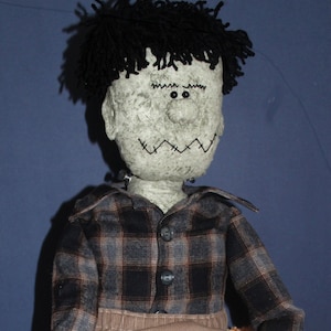 May include: A plush doll with a gray face, black hair, and black eyes. The doll is wearing a plaid shirt and brown pants. The doll is sitting on an orange fuzzy surface.