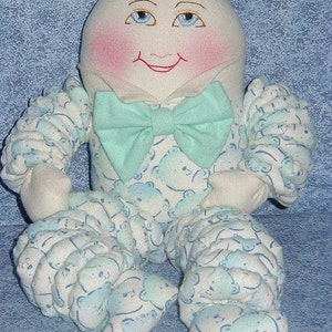 Könnte beinhalten: Eine handgefertigte Puppe mit einem weißen Gesicht, blauen Augen und rosigen Wangen. Die Puppe trägt ein blaues und weiß gemustertes Outfit mit einer hellblauen Schleife.