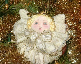 May include: A handmade Christmas angel ornament with a painted face, blonde hair, and a gold crown. The angel wears a cream-colored dress with gold accents and a large bow. The ornament is surrounded by gold tinsel and Christmas tree branches.