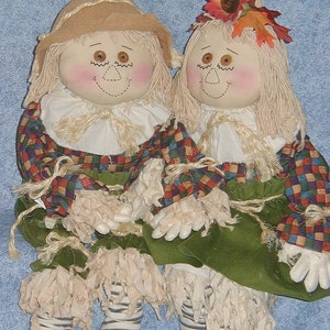 May include: Two handmade scarecrow dolls. One wears a tan hat, the other a floral headpiece. Both have yarn hair, rosy cheeks, and stitched facial features. They wear plaid shirts, green skirts, and striped socks with beige fringe.