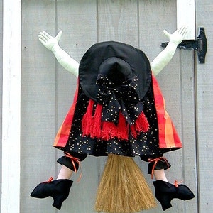May include: A Halloween decoration depicting a witch's body, arms, and legs. The witch is dressed in a black hat with gold stars, a black dress with gold stars and red accents, and black shoes. A broom is attached to the witch's body.