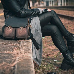 May include: A black and brown leather satchel bag with two front pockets and a top flap, secured with metal clasps. The bag is worn with black leather pants and boots. The image is taken outdoors.