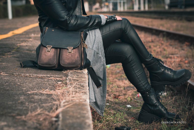 May include: A black and brown leather satchel bag with two front pockets and a top flap, secured with metal clasps. The bag is worn with black leather pants and boots. The image is taken outdoors.
