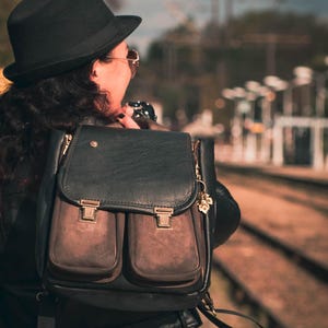 May include: Black leather backpack with two front pockets and gold-colored clasps. The backpack has a flap closure and a small gold charm. The bag is worn over the shoulder.