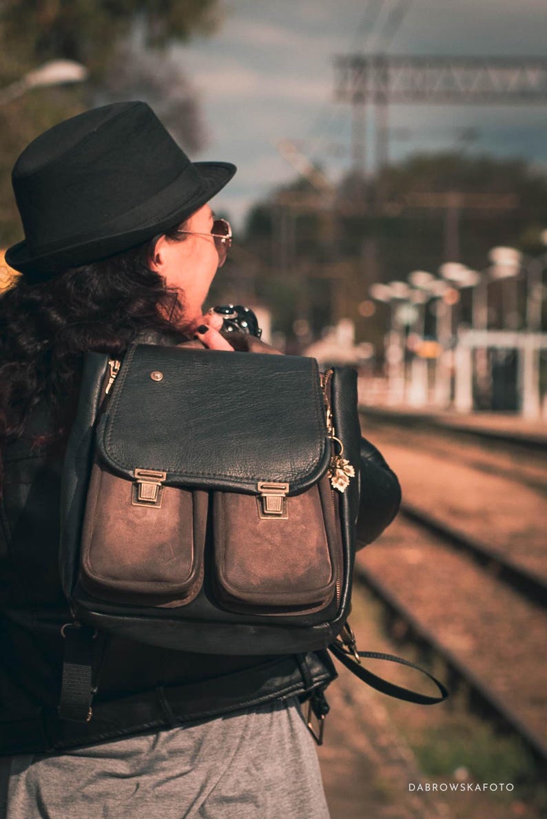 May include: Black leather backpack with two front pockets and gold-colored clasps. The backpack has a flap closure and a small gold charm. The bag is worn over the shoulder.