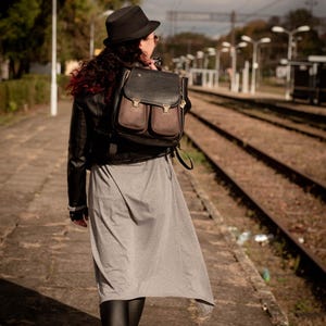 May include: A black and brown leather backpack with two front pockets is worn over the shoulder. The person is wearing a black hat, jacket, and boots, and a gray dress. The photo is taken at a train station.