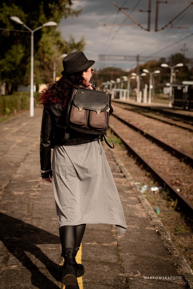 May include: A black and brown leather backpack with two front pockets is worn over the shoulder. The person is wearing a black hat, jacket, and boots, and a gray dress. The photo is taken at a train station.
