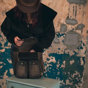 May include: A brown leather satchel with two front pockets and gold-colored hardware sits on a light blue dresser. The satchel has a black top and is open, revealing its interior. The background is a distressed wall.