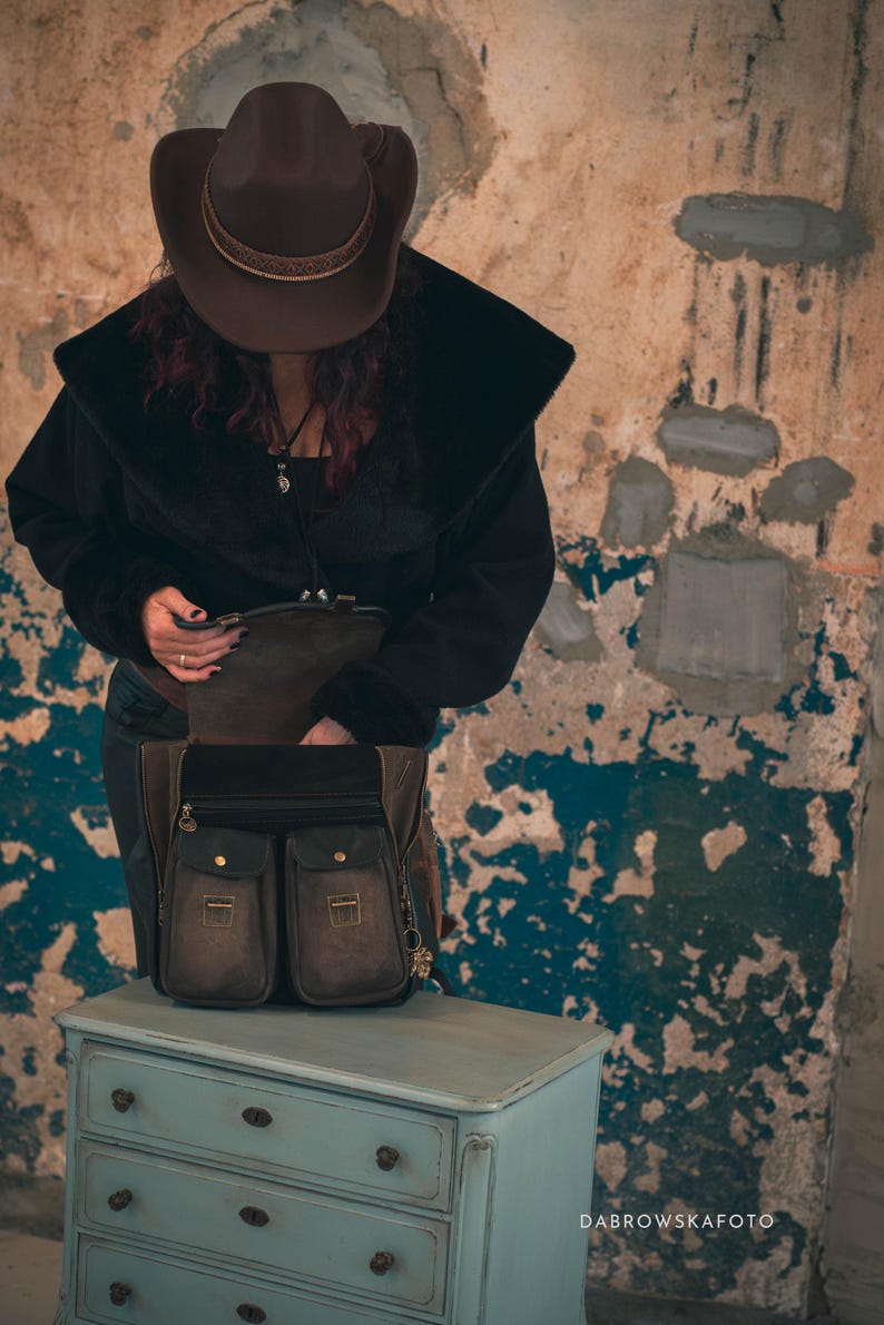 May include: A brown leather satchel with two front pockets and gold-colored hardware sits on a light blue dresser. The satchel has a black top and is open, revealing its interior. The background is a distressed wall.