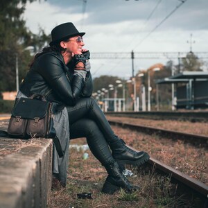 May include: A person wearing a black hat, sunglasses, and a black leather jacket sits near train tracks. A black leather backpack with brown accents rests nearby. The scene is set outdoors on a cloudy day.
