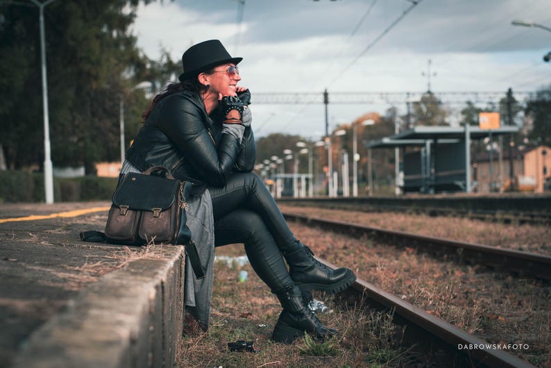 May include: A person wearing a black hat, sunglasses, and a black leather jacket sits near train tracks. A black leather backpack with brown accents rests nearby. The scene is set outdoors on a cloudy day.