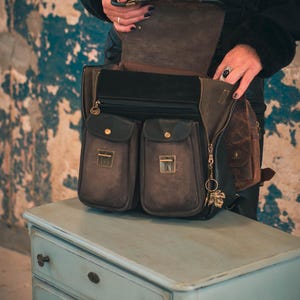 May include: A brown leather satchel with multiple pockets and a zippered main compartment. The bag has a flap closure with a gold-toned clasp and a decorative charm. The bag is sitting on a light blue dresser.