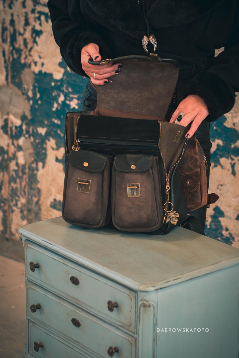 May include: A brown leather satchel with multiple pockets and a zippered main compartment. The bag has a flap closure with a gold-toned clasp and a decorative charm. The bag is sitting on a light blue dresser.