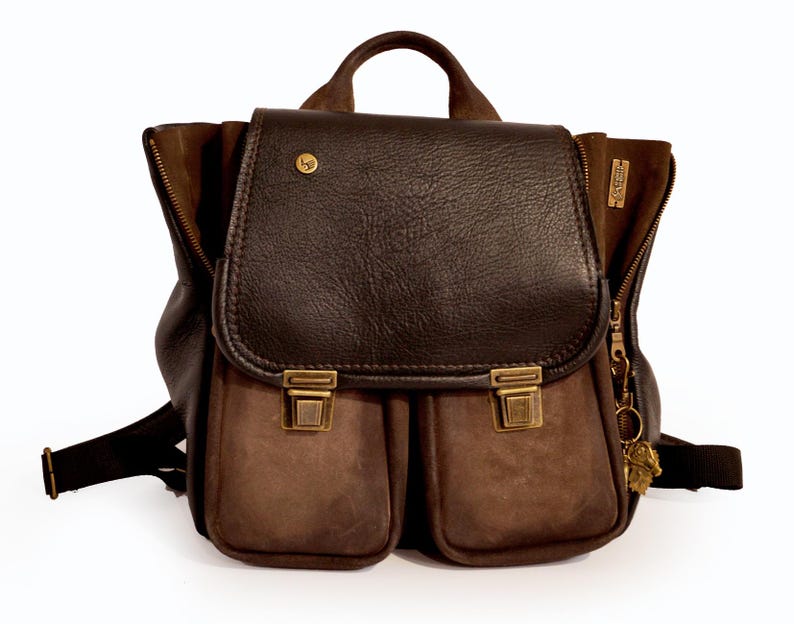 May include: A brown leather backpack with a flap closure and two front pockets. The backpack features a top handle, adjustable straps, and brass-colored hardware. The bag has a vintage aesthetic.