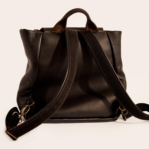 May include: A dark brown leather backpack with adjustable shoulder straps and a top handle. The bag has a flap closure and brass-colored hardware. The leather has a textured appearance.