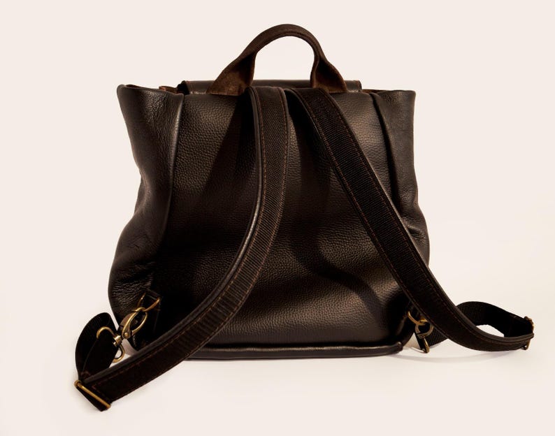 May include: A dark brown leather backpack with adjustable shoulder straps and a top handle. The bag has a flap closure and brass-colored hardware. The leather has a textured appearance.