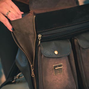 May include: A close-up of a brown and black leather bag with multiple pockets and a zipper closure. The bag has a flap with a gold-colored clasp and a small pocket with a gold button. The bag is open, revealing the interior.