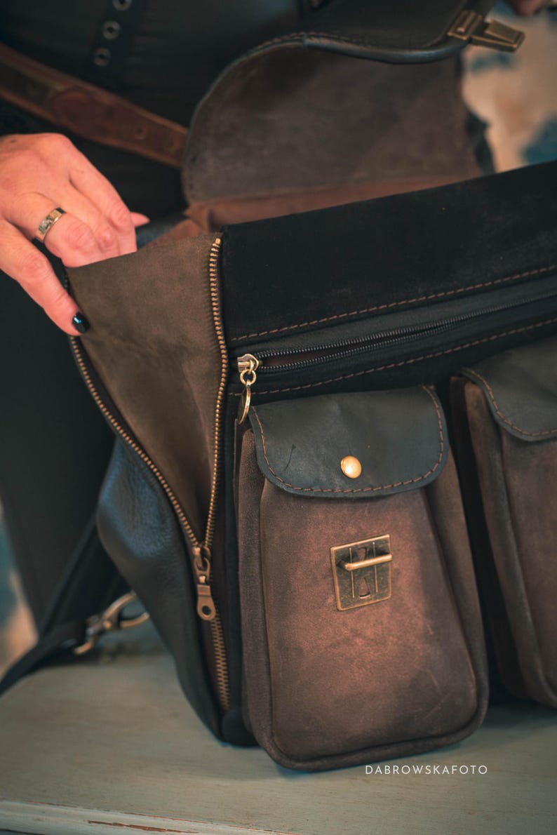 May include: A close-up of a brown and black leather bag with multiple pockets and a zipper closure. The bag has a flap with a gold-colored clasp and a small pocket with a gold button. The bag is open, revealing the interior.