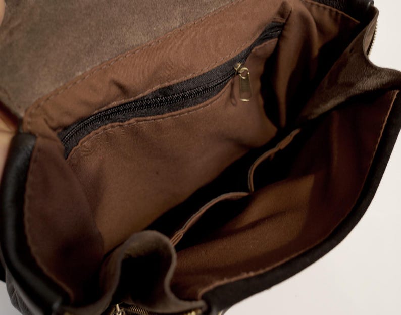 May include: Open view of a black leather bag's interior, revealing a brown fabric lining and multiple pockets. A black zipper runs across the top, and a gold-colored zipper pull is visible. The bag's interior is well-lit, showcasing the details.