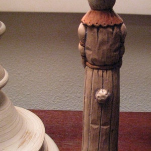 Pu&ograve; includere: Una statuetta di coniglio di legno che indossa un cappello e una lunga veste marrone. La statuetta &egrave; in piedi su una base di legno.