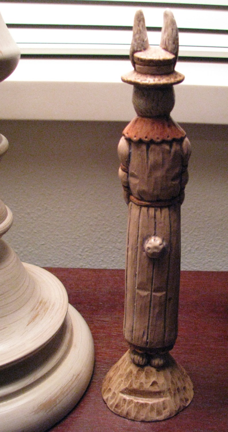 Pu&ograve; includere: Una statuetta di coniglio di legno che indossa un cappello e una lunga veste marrone. La statuetta &egrave; in piedi su una base di legno.
