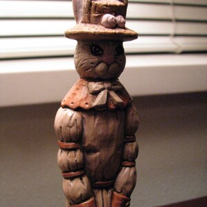 Pu&ograve; includere: Una statuetta in ceramica di un coniglio che indossa un cappello e un vestito. Il coniglio ha la pelliccia marrone e un naso rosa. Il cappello &egrave; marrone con un fiore rosa in cima. Il vestito &egrave; marrone con un colletto bianco e un fiocco bianco.