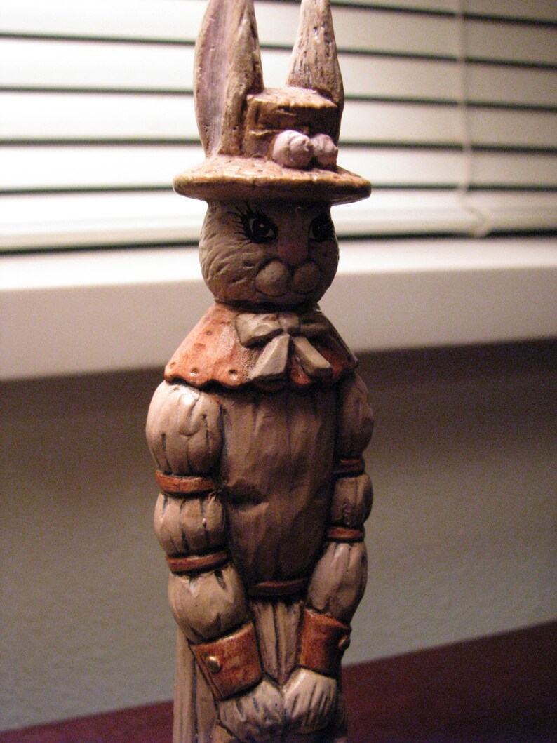 Pu&ograve; includere: Una statuetta in ceramica di un coniglio che indossa un cappello e un vestito. Il coniglio ha la pelliccia marrone e un naso rosa. Il cappello &egrave; marrone con un fiore rosa in cima. Il vestito &egrave; marrone con un colletto bianco e un fiocco bianco.