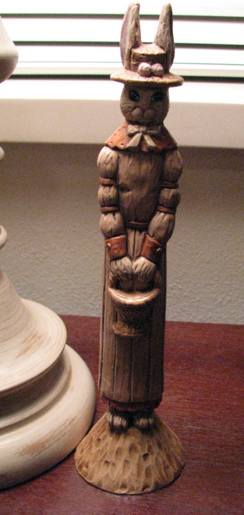 Pu&ograve; includere: Una statuetta in ceramica di un coniglio che indossa un cappello e un vestito. Il coniglio tiene un secchio nelle sue mani. La statuetta &egrave; dipinta in tonalit&agrave; di marrone e bianco.