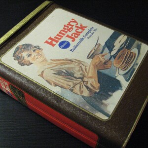 May include: A brown and gold box with a vintage design. The box features a woman pouring batter into a pan and the text "Hungry Jack Buttermilk Complete Pancake Mix".
