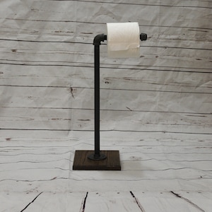 May include: Black metal toilet paper holder with a wooden base. The holder is mounted on a white wood background.