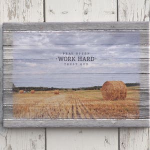 May include: A rustic wooden sign with a faded blue sky and field of hay bales. The sign reads "Pray Often, Work Hard, Trust God".