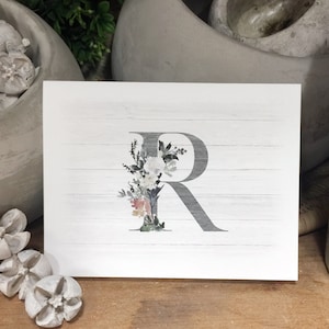 May include: A white card with a gray letter R decorated with watercolor flowers on a white woodgrain background.
