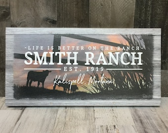 May include: A rustic wooden sign with a sunset background and silhouettes of cattle. The sign reads "Life is better on the ranch, Smith Ranch, Est. 1919, Kalispell, Montana."