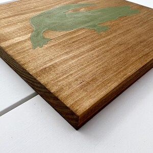 Puede incluir: Un letrero de madera con una silueta verde de una rana. El letrero est&aacute; te&ntilde;ido de un color marr&oacute;n claro.
