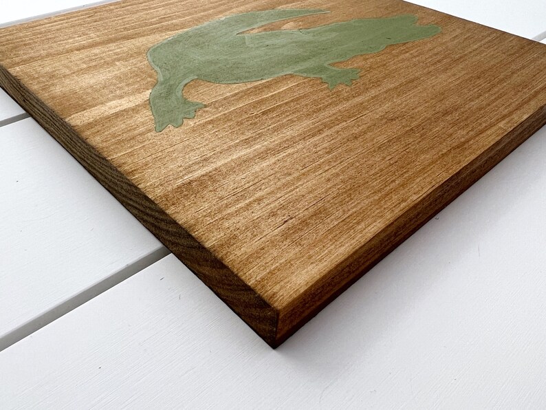 Puede incluir: Un letrero de madera con una silueta verde de una rana. El letrero est&aacute; te&ntilde;ido de un color marr&oacute;n claro.