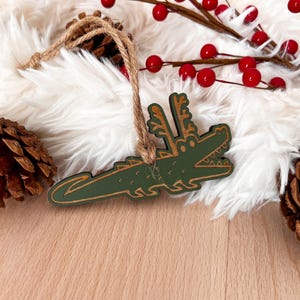 Puede incluir: Adorno navide&ntilde;o verde con forma de cocodrilo con contorno marr&oacute;n y una cuerda de yute para colgar. El adorno se coloca sobre un fondo de piel sint&eacute;tica blanca, pi&ntilde;as y bayas rojas.