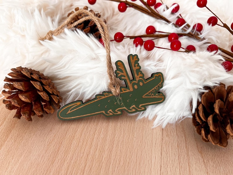 Puede incluir: Adorno navide&ntilde;o verde con forma de cocodrilo con contorno marr&oacute;n y una cuerda de yute para colgar. El adorno se coloca sobre un fondo de piel sint&eacute;tica blanca, pi&ntilde;as y bayas rojas.