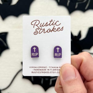 May include: A pair of purple RIP tombstone earrings. The earrings are on a white card with the text "Rustic Strokes". The earrings are held by a person's hand. The background has black bat cutouts.