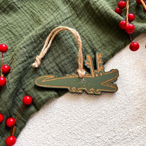 Puede incluir: Adorno de madera verde con forma de cocodrilo con una cuerda de yute para colgar. El cocodrilo tiene un dise&ntilde;o detallado con una boca sonriente y cuernos. Bayas rojas y tela verde en el fondo.