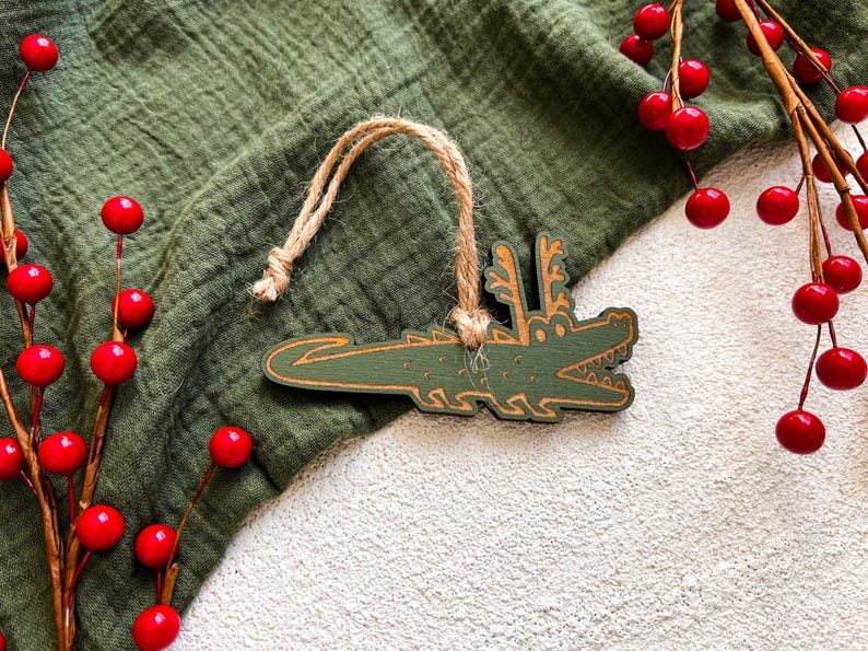 Puede incluir: Adorno de madera verde con forma de cocodrilo con una cuerda de yute para colgar. El cocodrilo tiene un dise&ntilde;o detallado con una boca sonriente y cuernos. Bayas rojas y tela verde en el fondo.