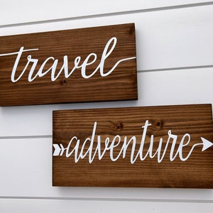 May include: Two wooden signs with white lettering. The top sign says "travel" and the bottom sign says "adventure" with an arrow pointing to the right.