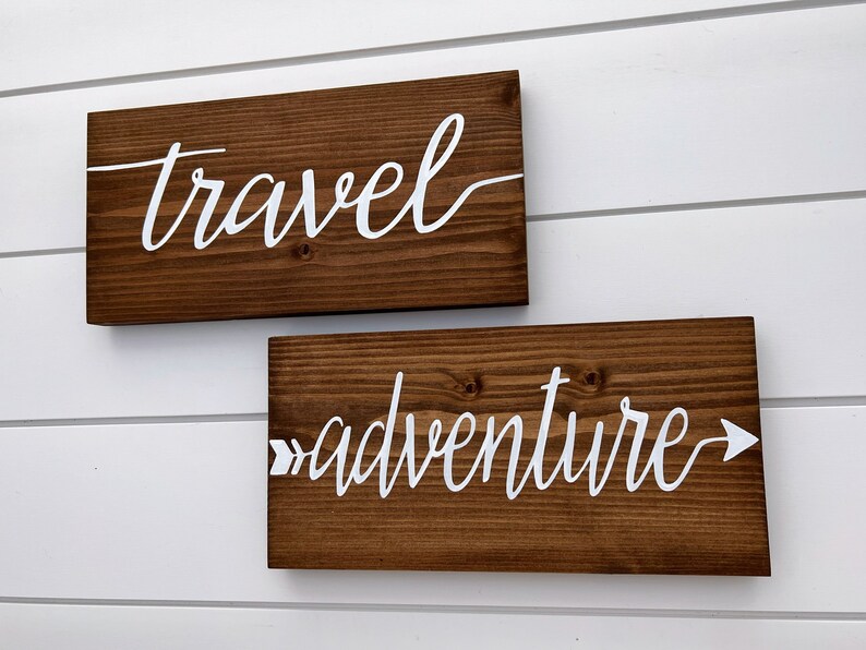 May include: Two wooden signs with white lettering. The top sign says "travel" and the bottom sign says "adventure" with an arrow pointing to the right.