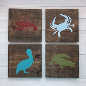 Puede incluir: Cuatro cuadrados de madera con siluetas pintadas de criaturas marinas. El cuadrado superior izquierdo tiene una langosta roja, el cuadrado superior derecho tiene un cangrejo azul, el cuadrado inferior izquierdo tiene un pel&iacute;cano azul verdoso, y el cuadrado inferior derecho tiene un cocodrilo verde.