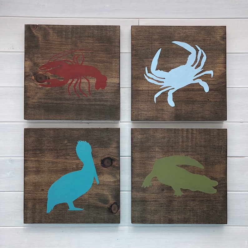 Puede incluir: Cuatro cuadrados de madera con siluetas pintadas de criaturas marinas. El cuadrado superior izquierdo tiene una langosta roja, el cuadrado superior derecho tiene un cangrejo azul, el cuadrado inferior izquierdo tiene un pel&iacute;cano azul verdoso, y el cuadrado inferior derecho tiene un cocodrilo verde.
