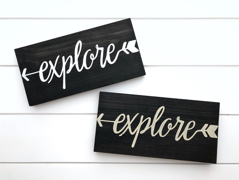 May include: Two black wooden signs with the word "explore" written in white and gold paint. Each sign has a white and gold arrow pointing to the left.